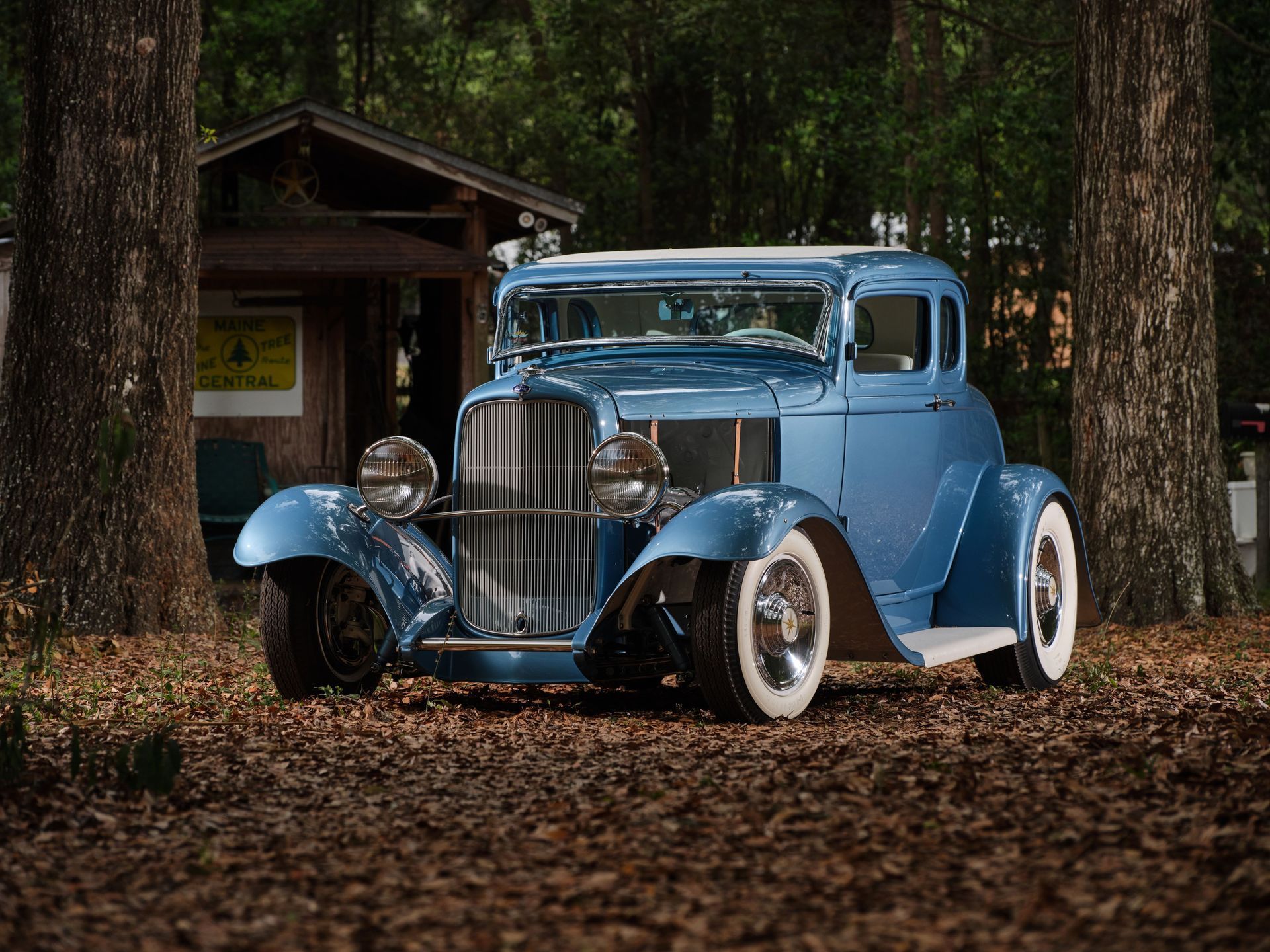 Blue vintage car parked on leaf-covered ground beside a small wooden cabin in the woods