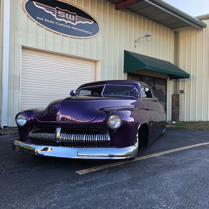 Purple classic car parked outside a tan metal building with a green awning and garage door