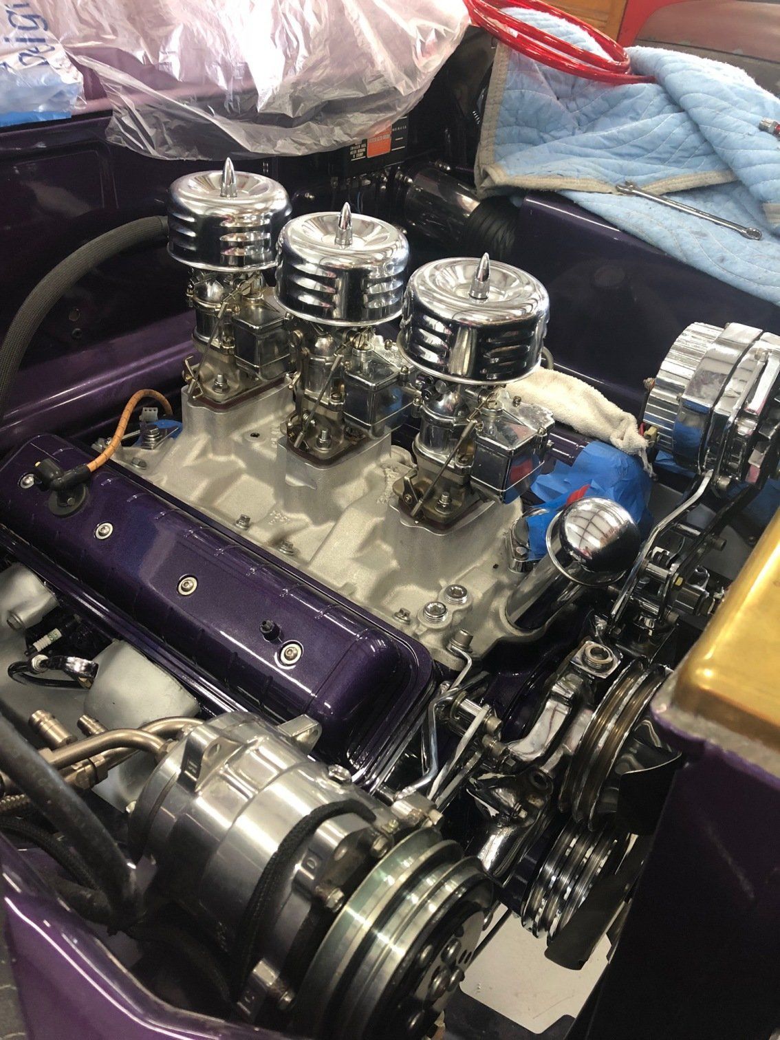 Custom car engine with three chrome air filters and purple valve covers in a garage.
