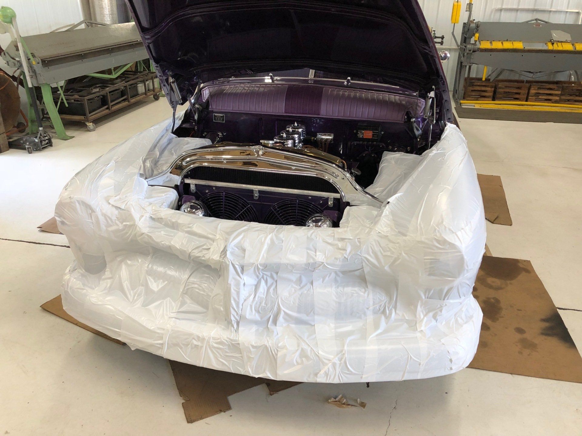 Car wrapped in white protective plastic inside a workshop, with hood open and front disassembled