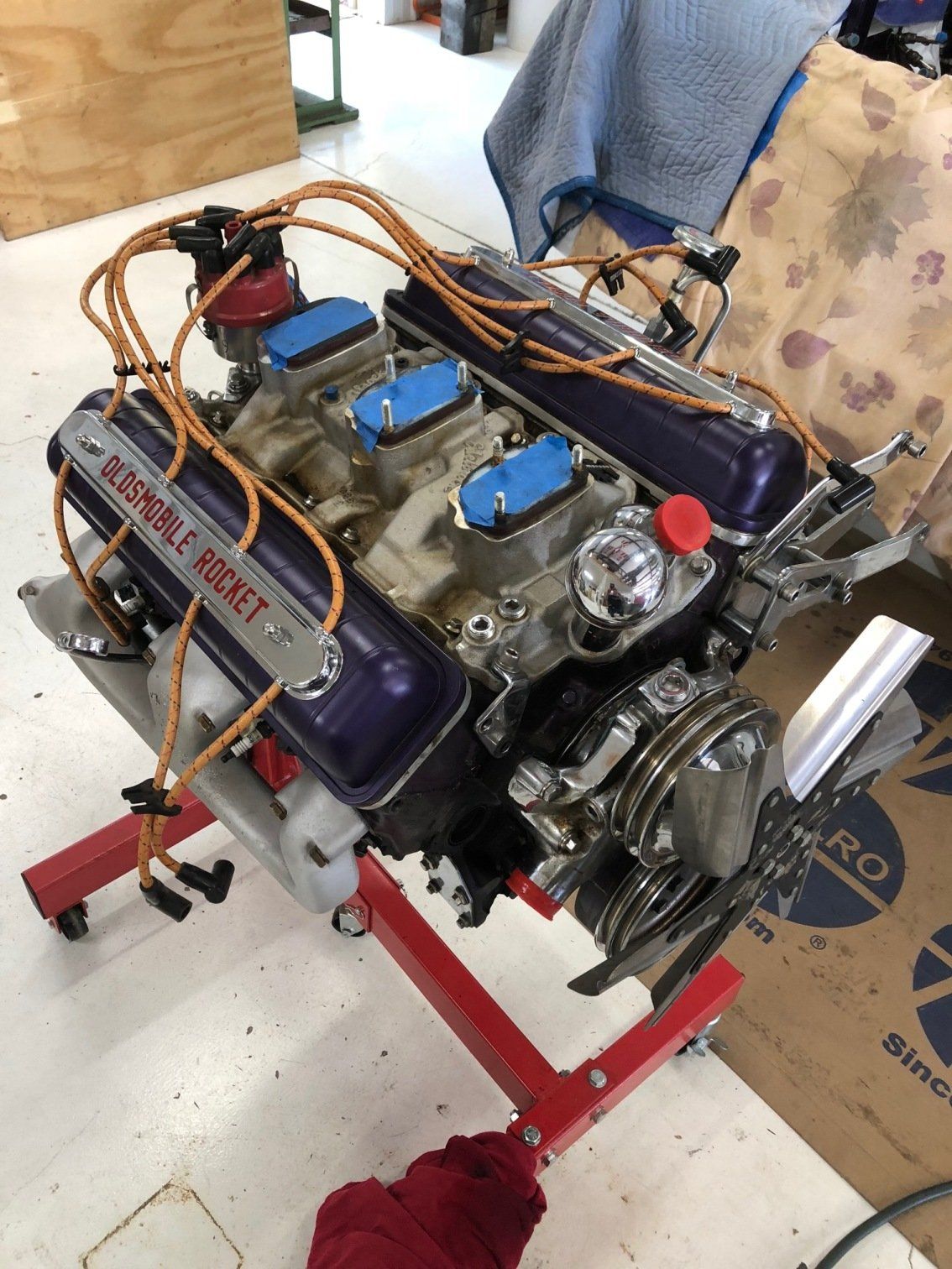 V8 engine mounted on a red stand, with blue valve covers and visible hoses in a workshop.
