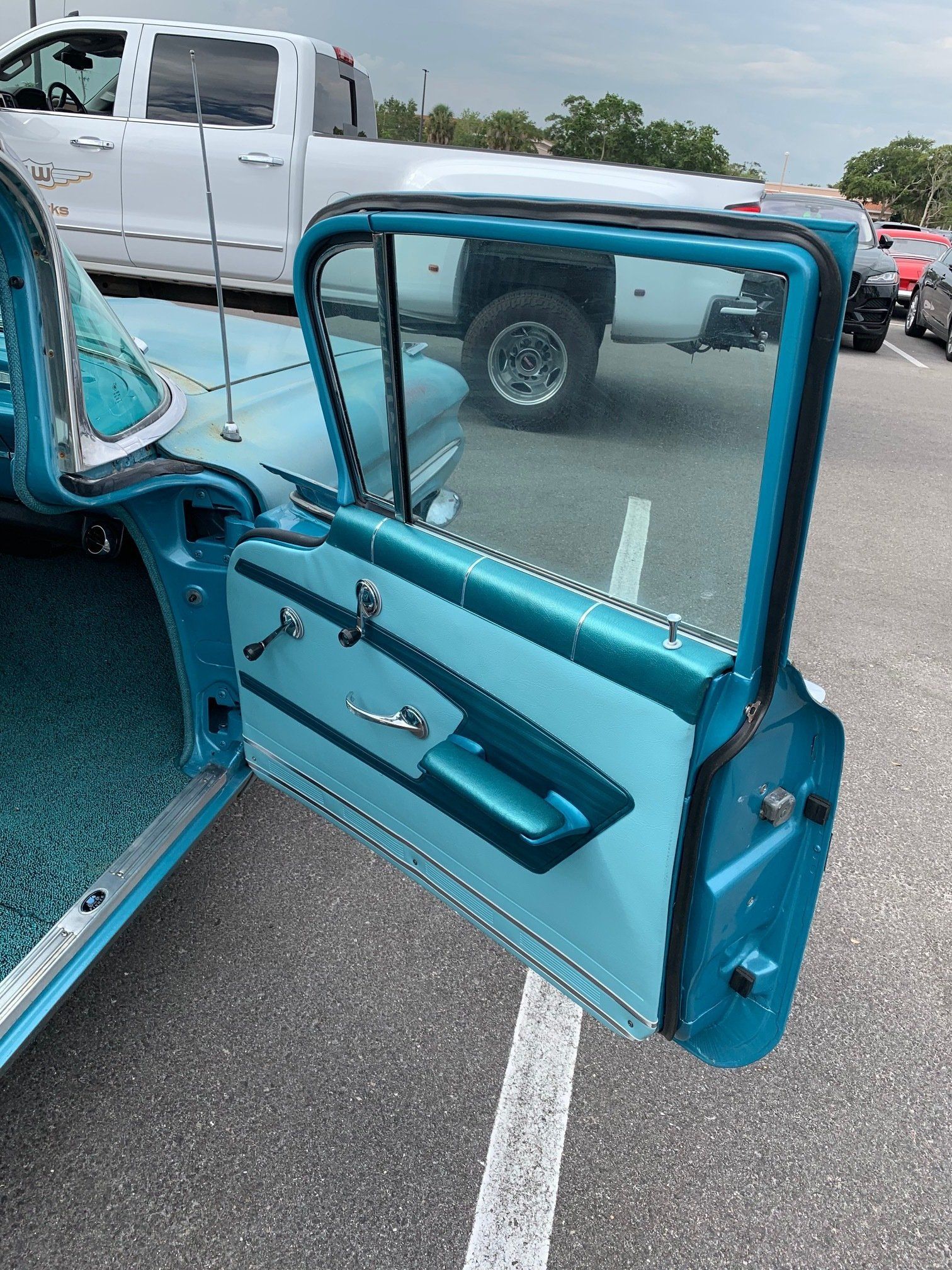 Open car door with turquoise interior in a parking lot, with a white truck in the background