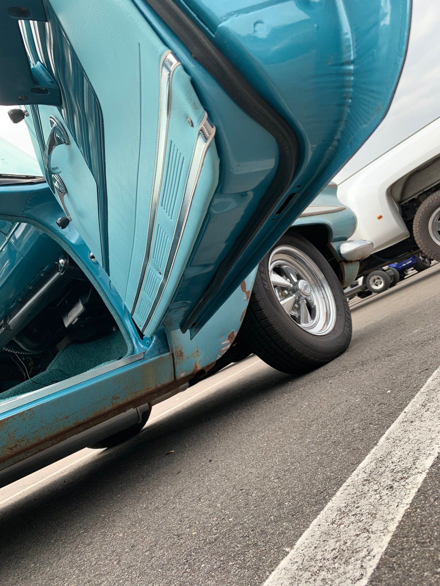 Turquoise vintage car body with open hood, parked low in a lot near a white vehicle and road markings.