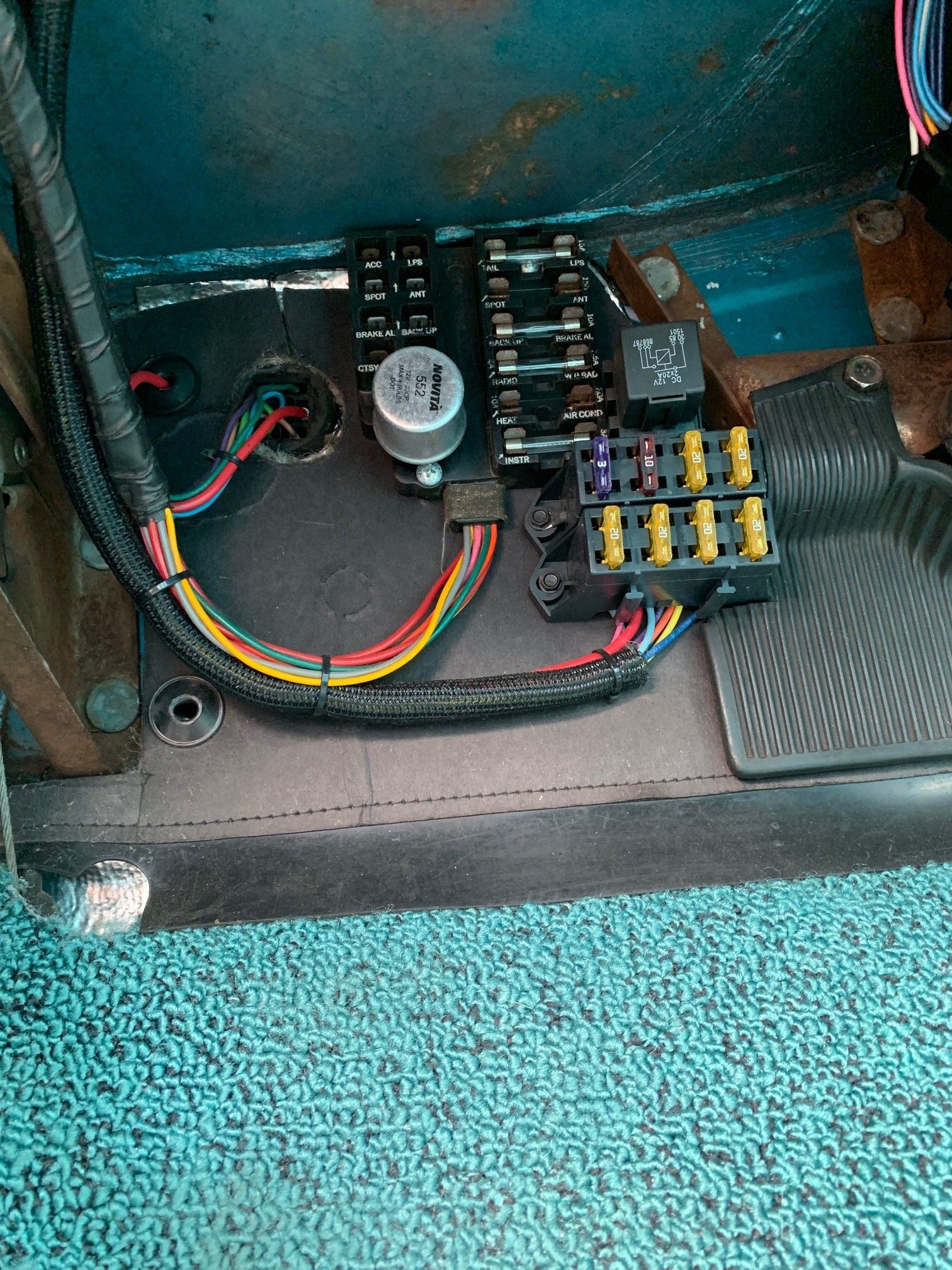 Car fuse box with exposed wiring under the dashboard, next to a vented panel and floor mat.