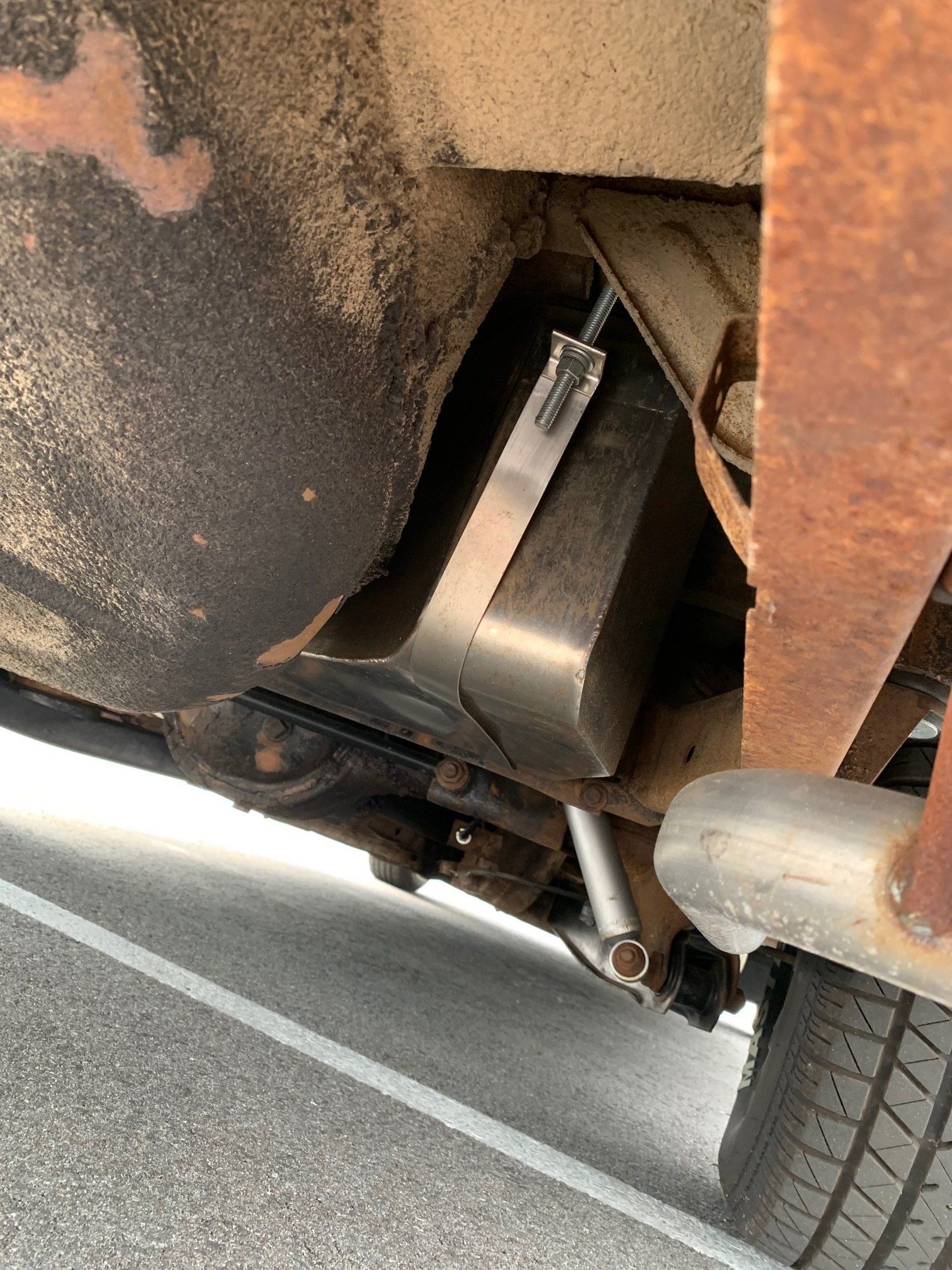 Undercarriage of a rusted vehicle showing a metal fuel tank strapped in place and a rear tire.