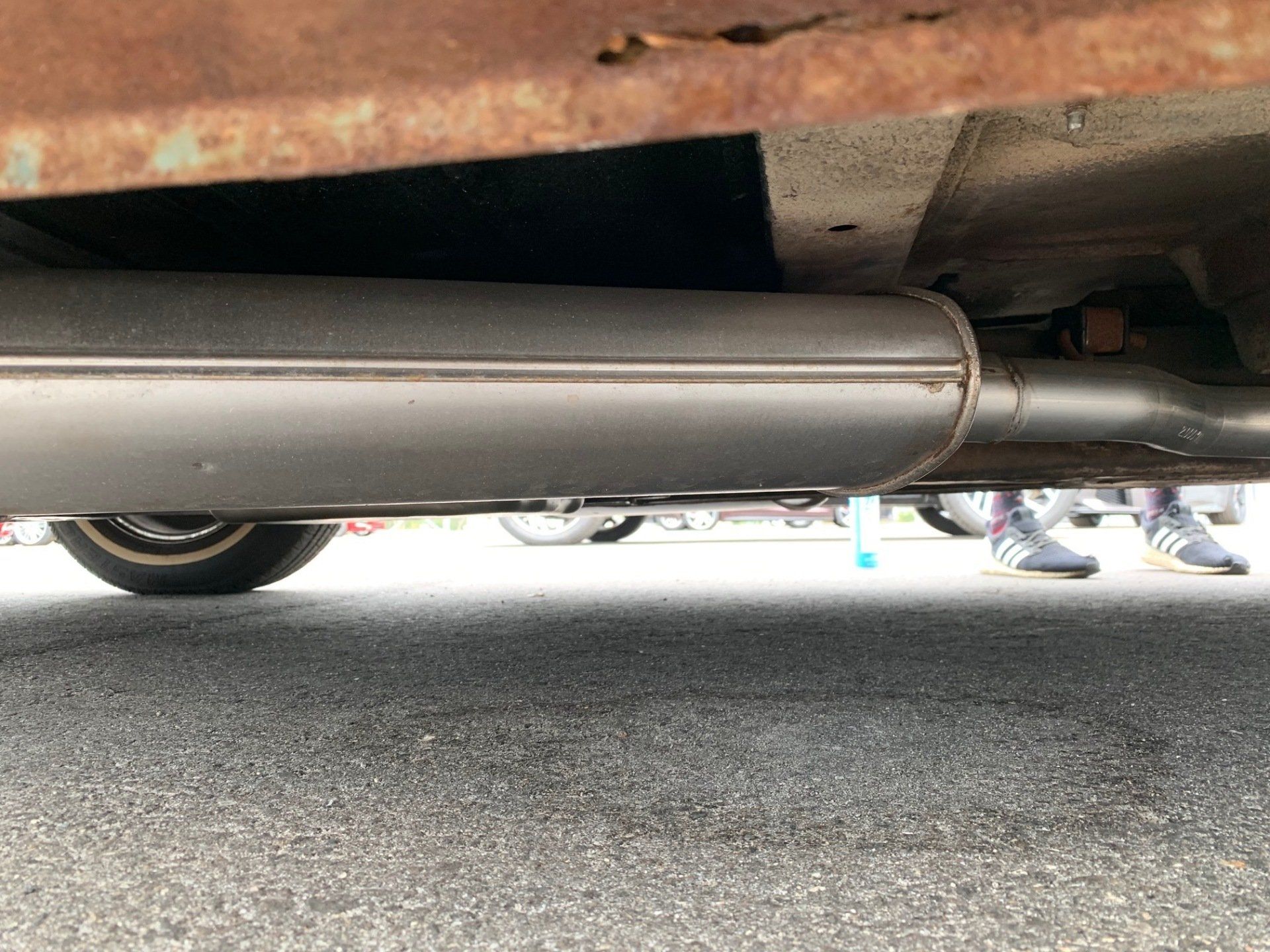 Under a car’s rear exhaust pipe, with a wheel and people’s shoes visible in the background