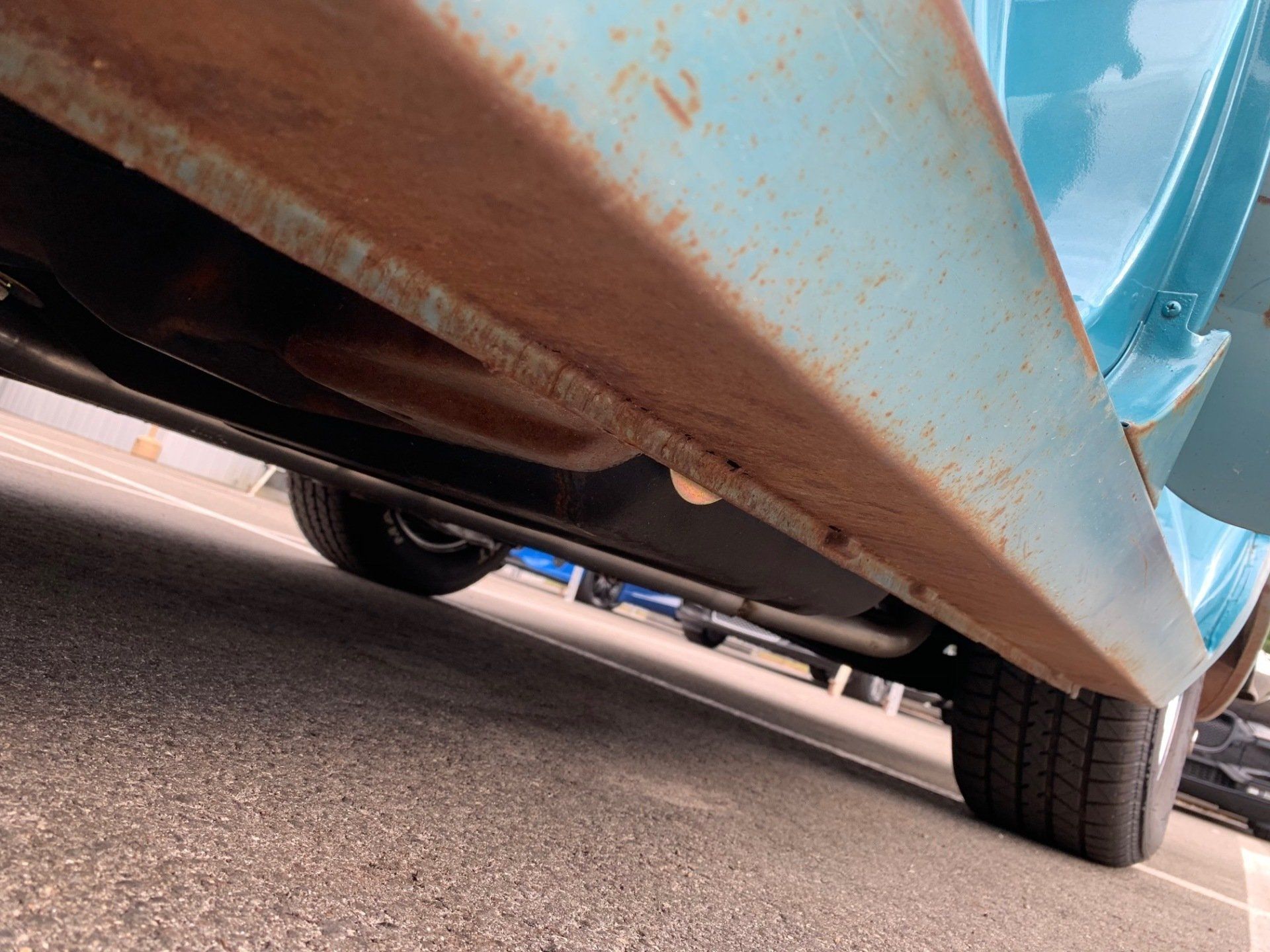 Rusty blue car chassis viewed from below, with wheel and road visible underneath.