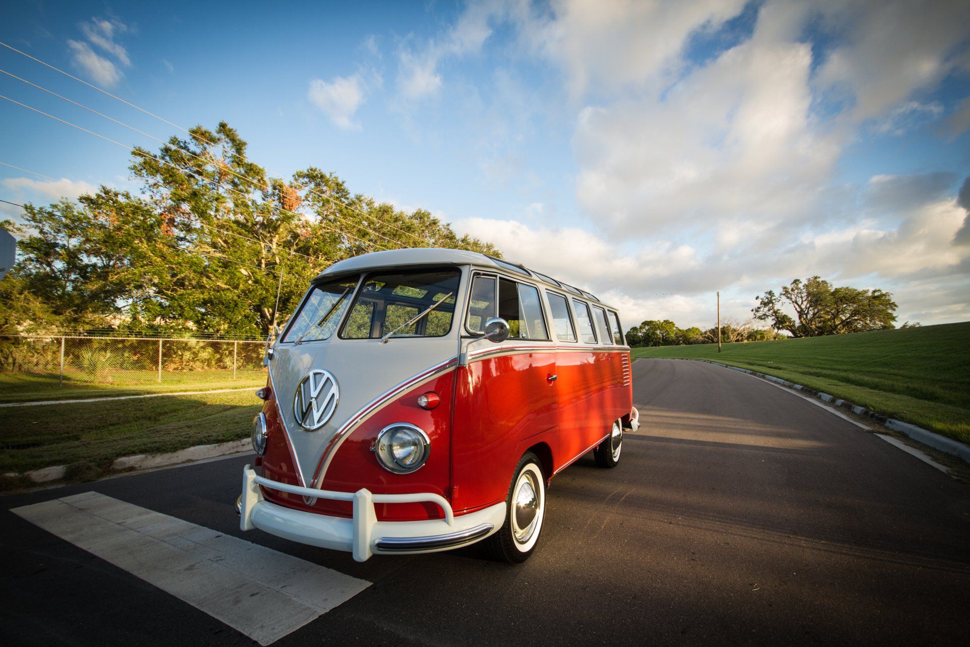 Red and white vintage Volkswagen van driving on a curved road under a blue sky with trees.
