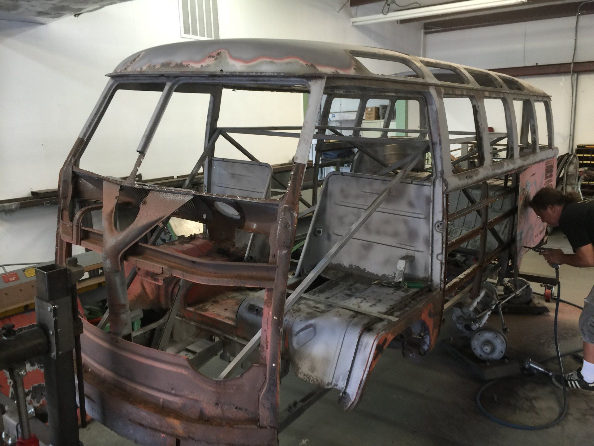 Rusty stripped-down van shell in a workshop, with exposed frame and a person working beside it