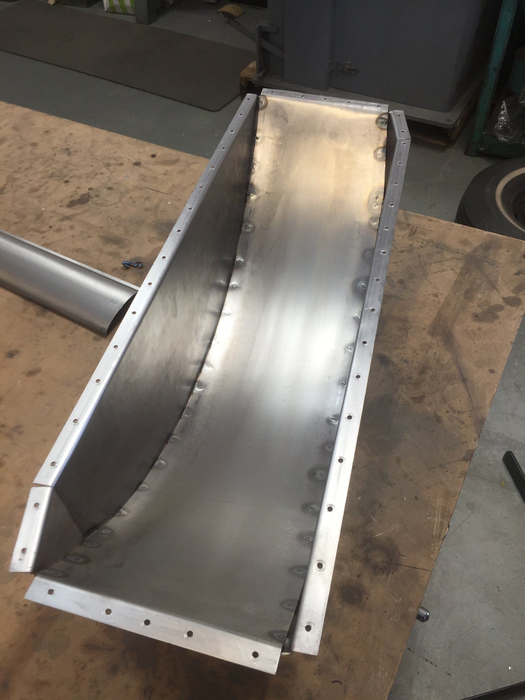 Metal chute with bolted white sides and a shiny curved interior on a workshop floor.