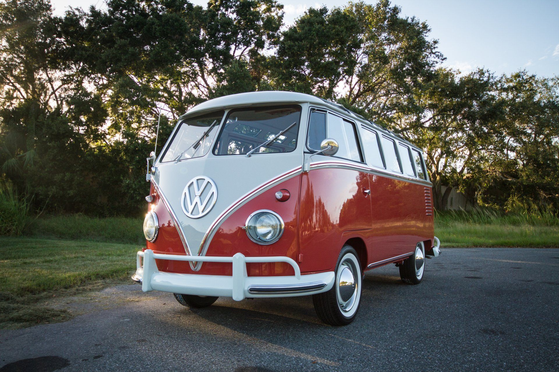 Red and white vintage Volkswagen van parked on a road at sunset, with trees in the background