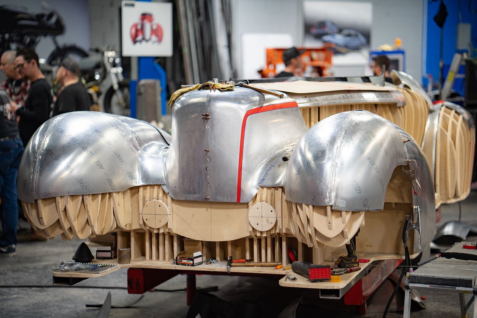 Metal car body prototype in workshop, with silver curved panels and exposed wooden supports on a wheeled cart