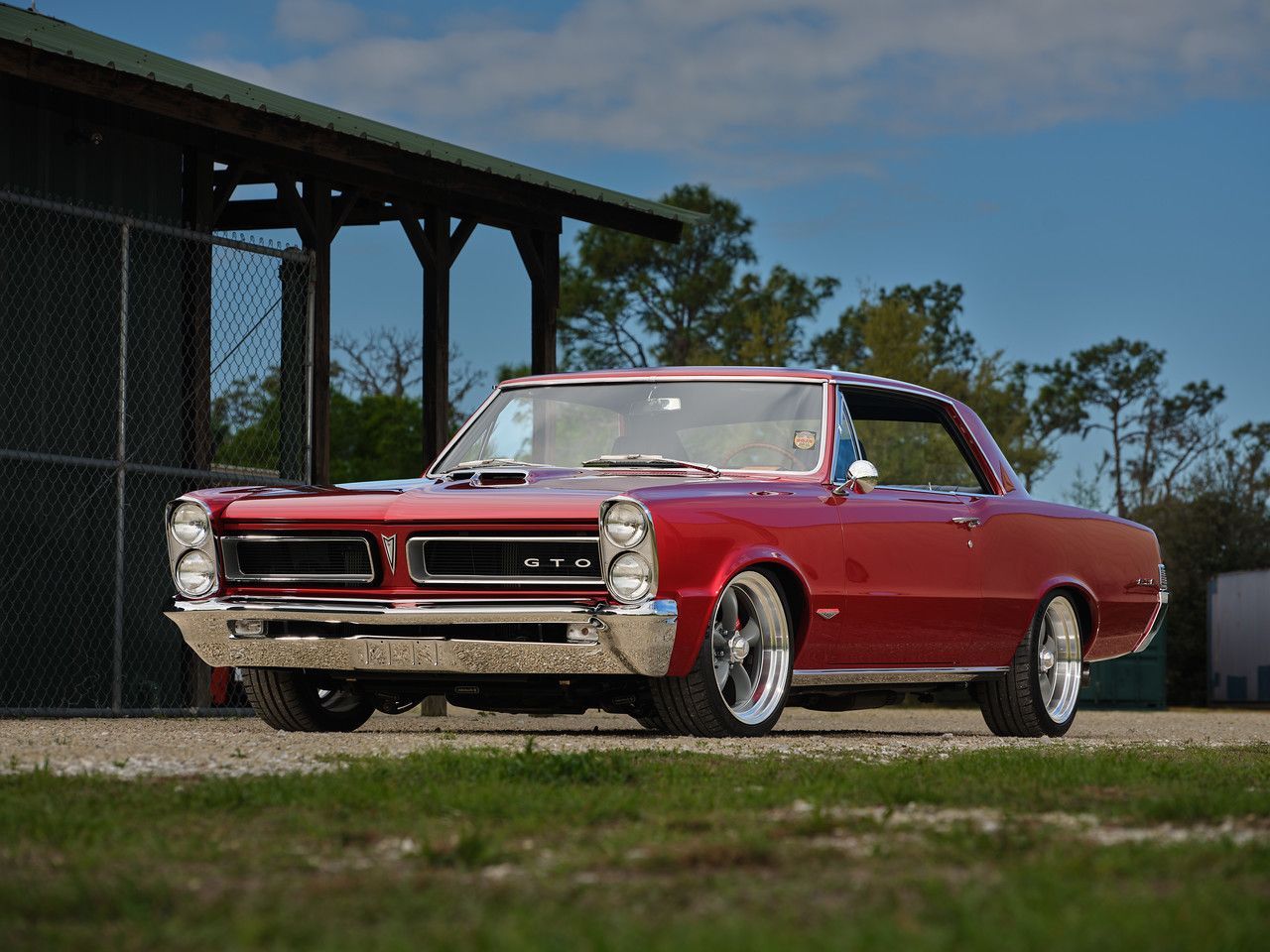 Red classic convertible parked on gravel beside a chain-link fence and shed under a blue sky.
