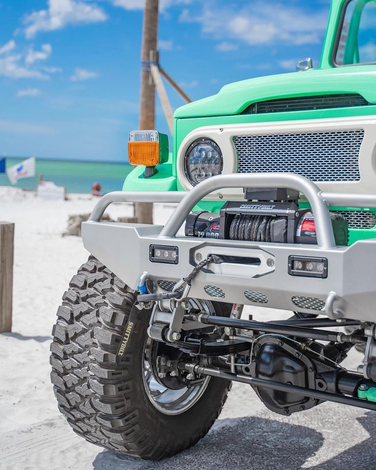 Custom green off-road truck front end with winch and large tire parked near a beach