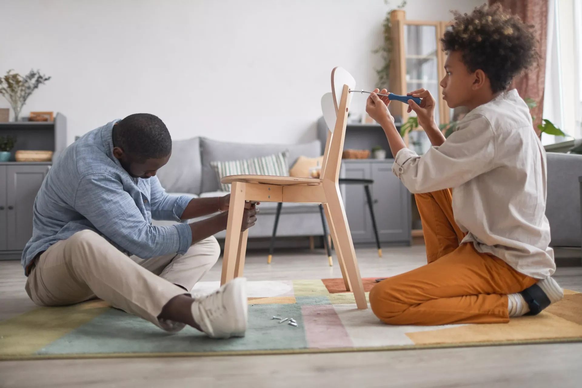 Father and child assembling a wooden chair on a rug indoors.
