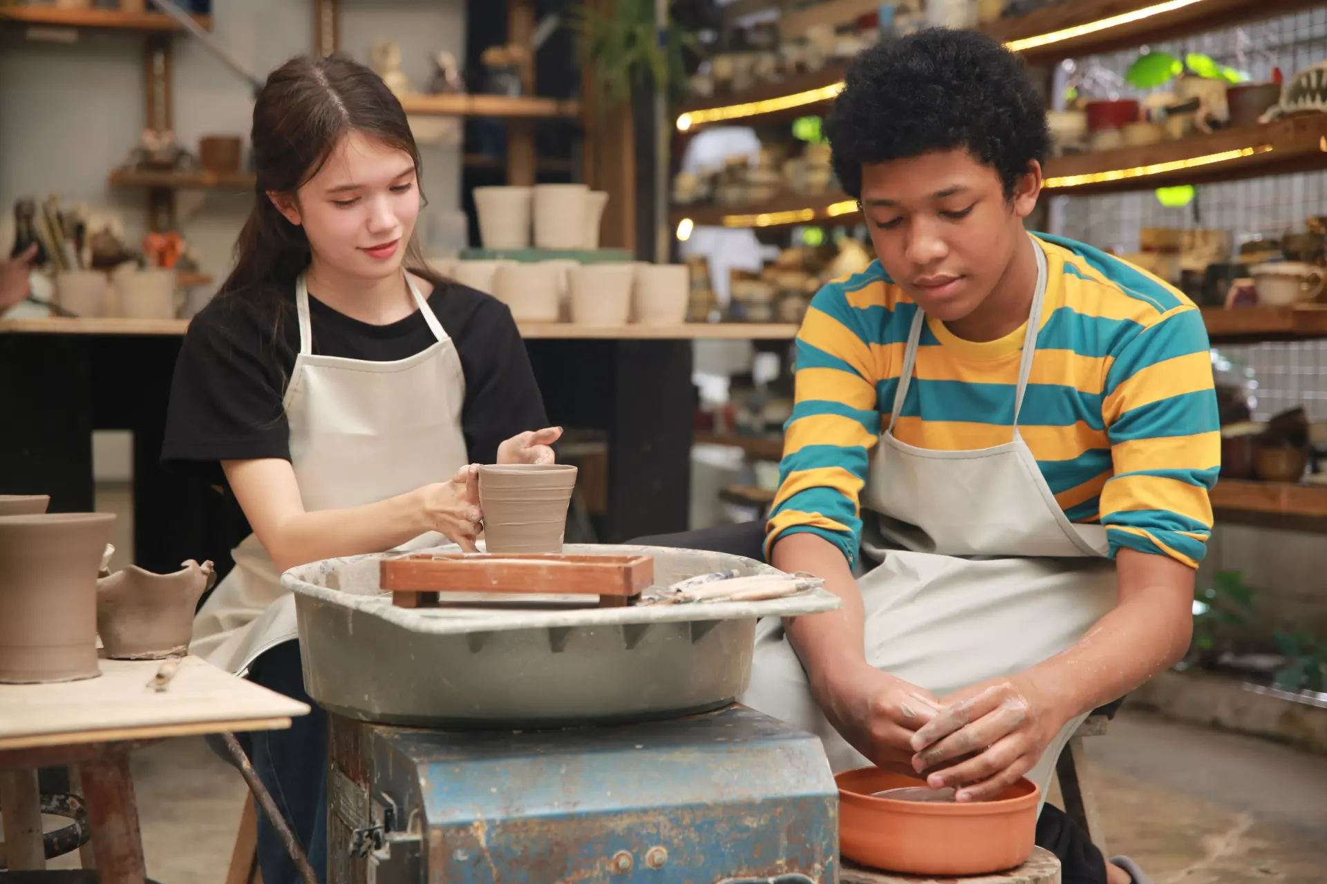Two people in aprons working with clay on a pottery wheel in a workshop.