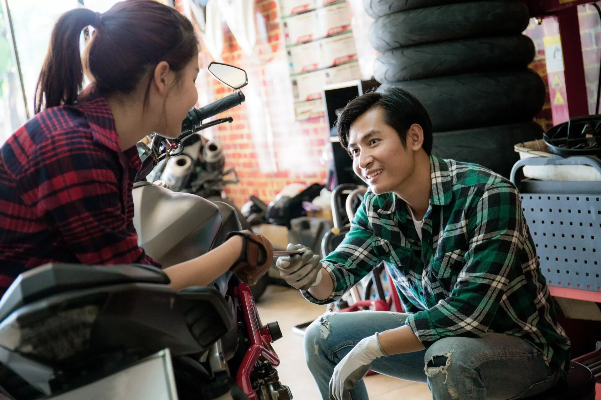 A person wearing a green plaid shirt smiles at another as they work on a motorcycle in a garage.