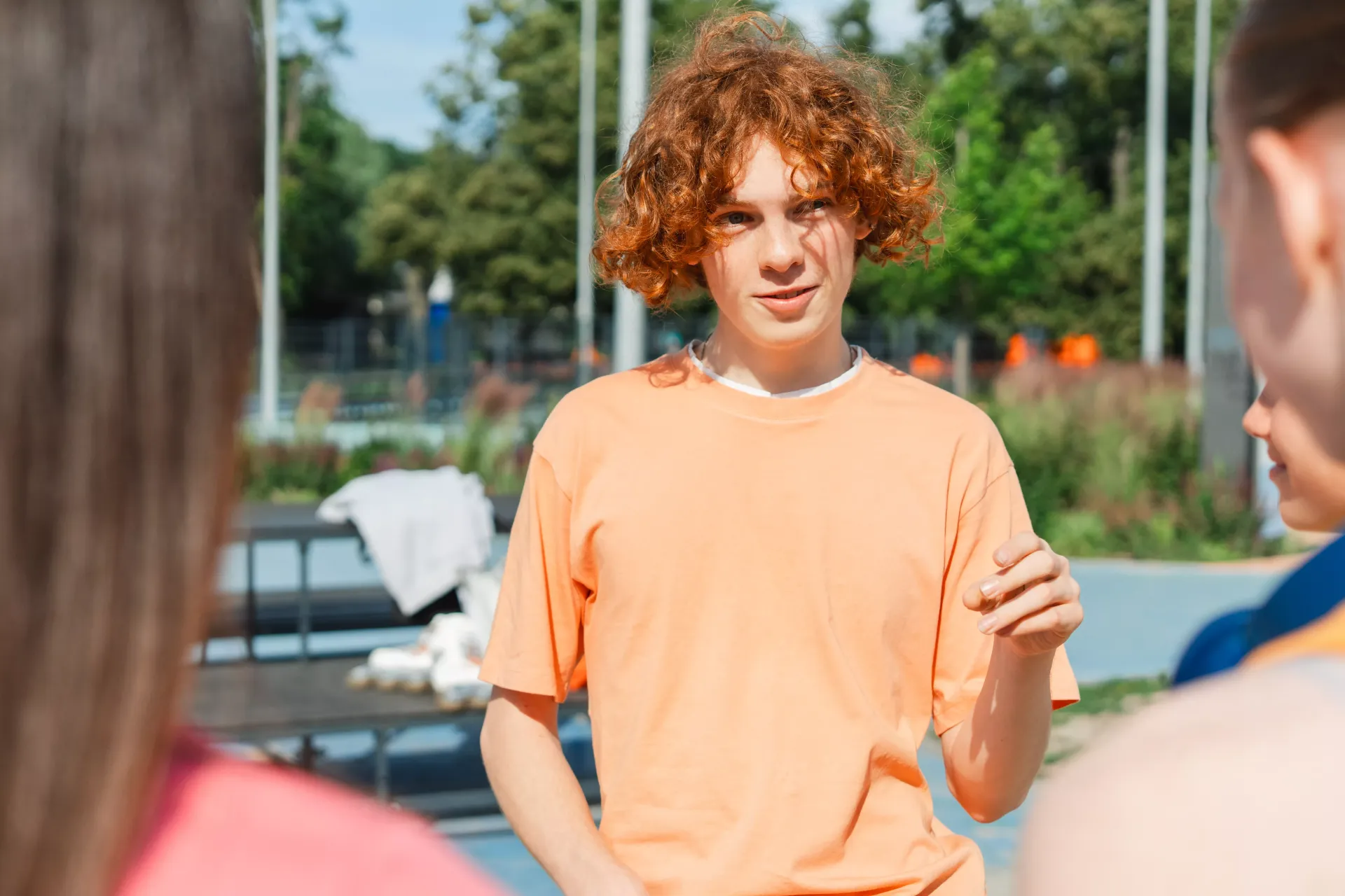 Teenager with red curly hair, orange shirt, talking with two girls outside on a sunny day.