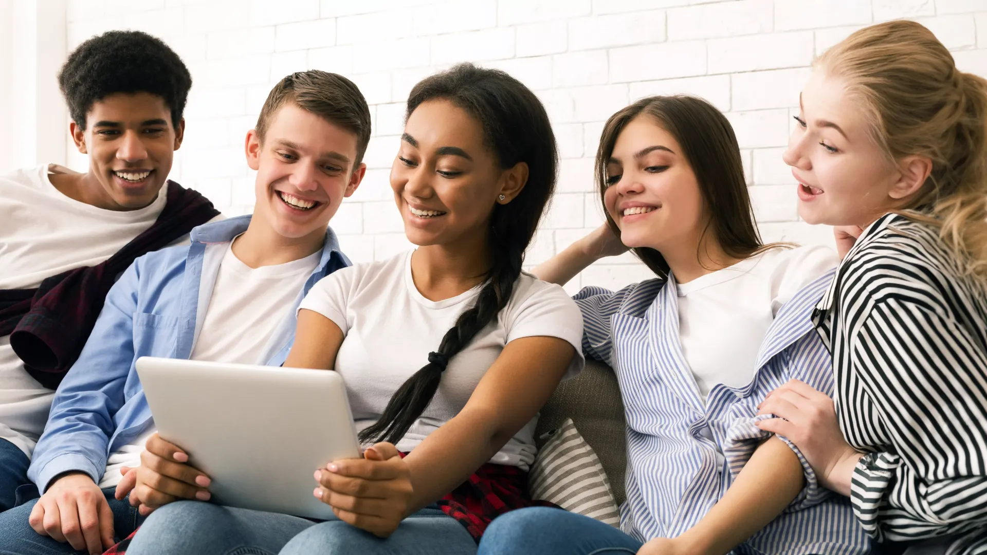 Five diverse young adults laugh while looking at a tablet together on a couch.
