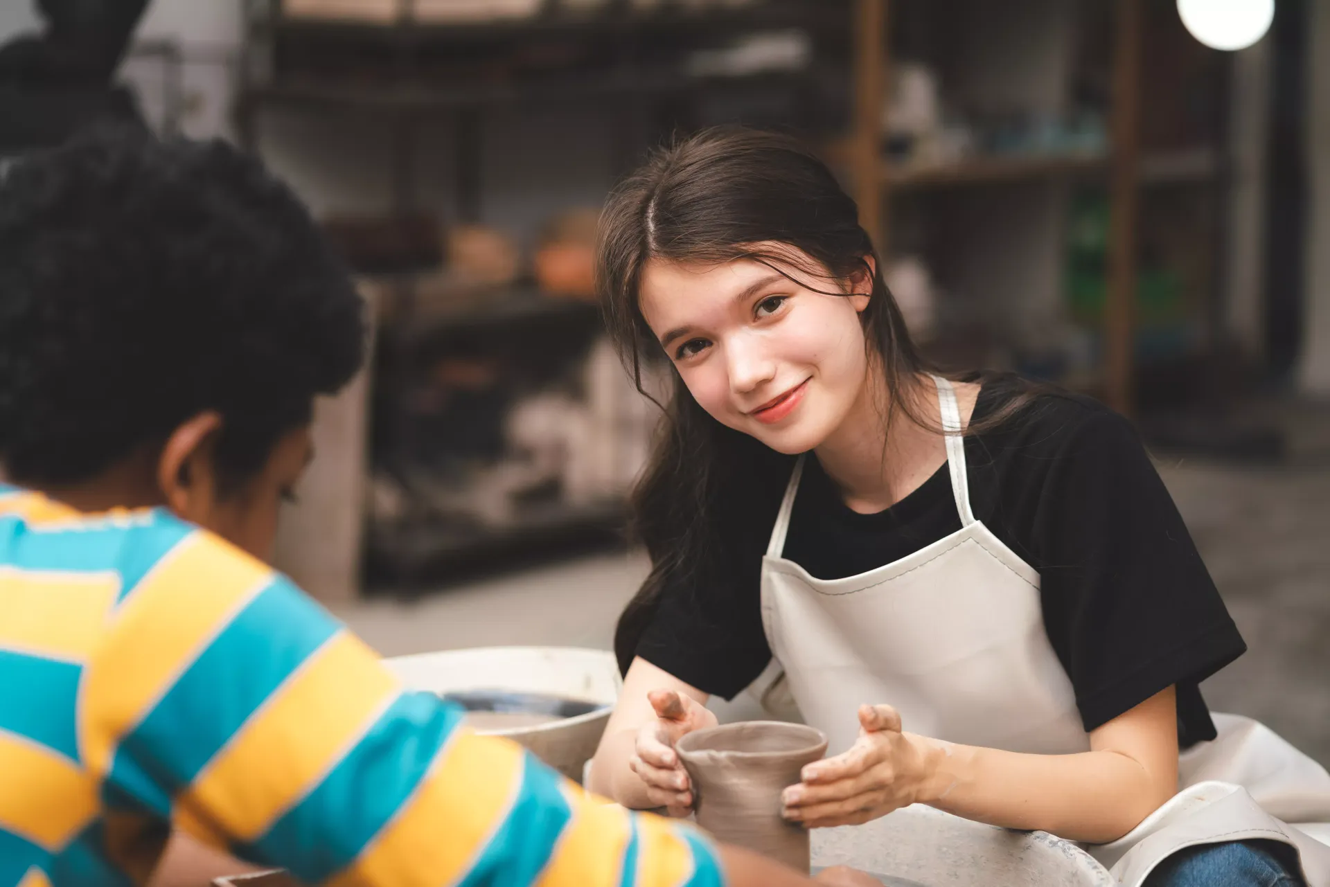 Woman and boy in a pottery class. Woman smiles, wearing an apron. Boy works on pottery at a wheel.