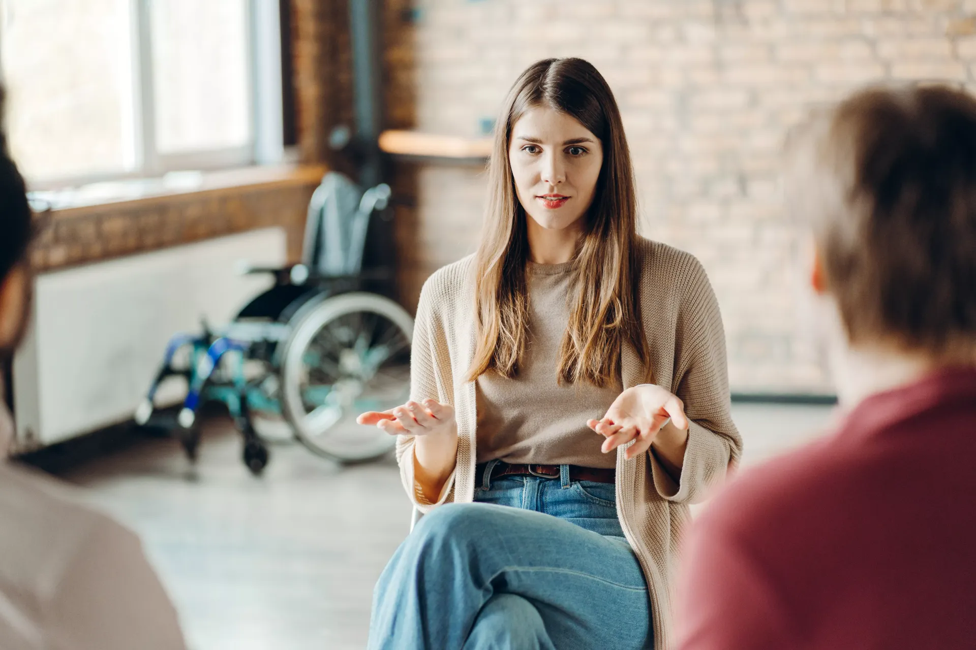 Woman in a group setting gestures while speaking; wheelchair visible in the background.