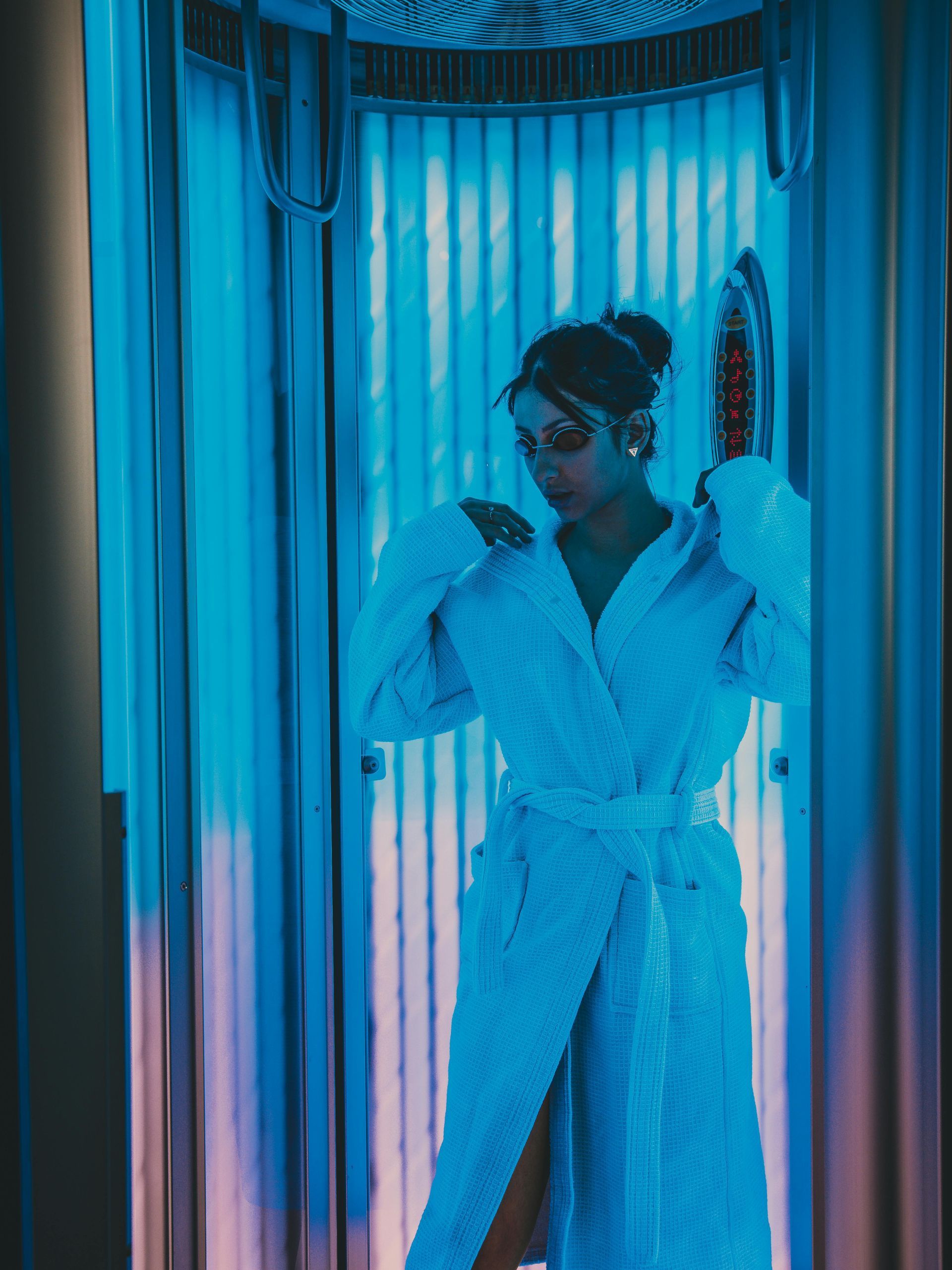 Person in a white robe standing in a blue-lit room, adjusting their hair by a mirrored wall.