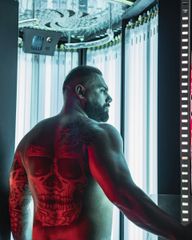 Bare-chested man in a futuristic chamber, lit blue and red, showing a glowing skull tattoo on his back