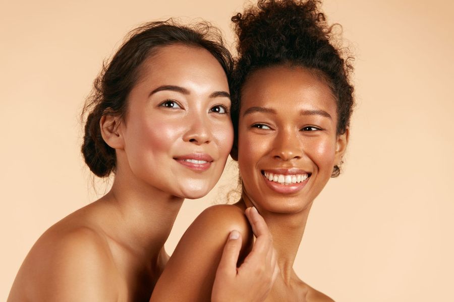 Two smiling people posing against a soft peach background, one resting a hand on the other’s shoulder.