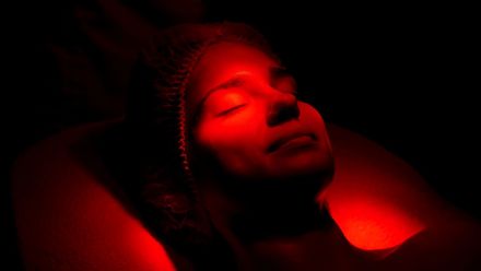 Person lying under red light with eyes closed, face partially illuminated in a dark setting