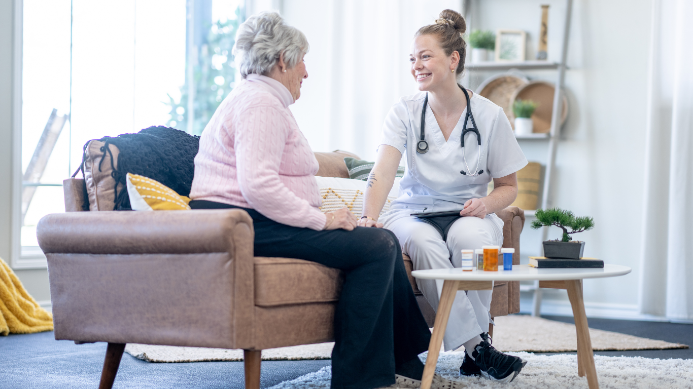 A nurse is talking to an elderly woman sitting on a couch.