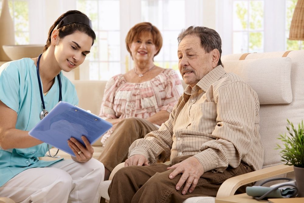 A nurse is talking to an elderly man sitting on a couch.