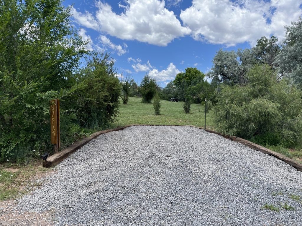 Gravel road leading into a green, grassy field with trees under a blue sky with clouds.