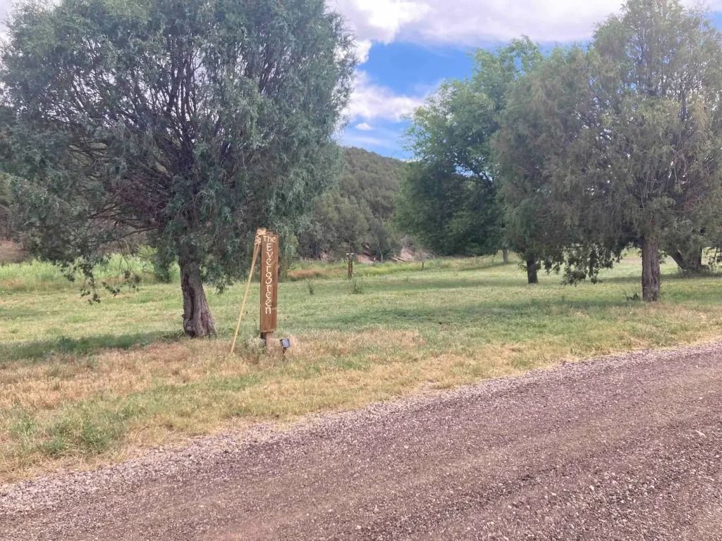 Gravel road leads to a grassy area with trees and a sign. The sky is partly cloudy.