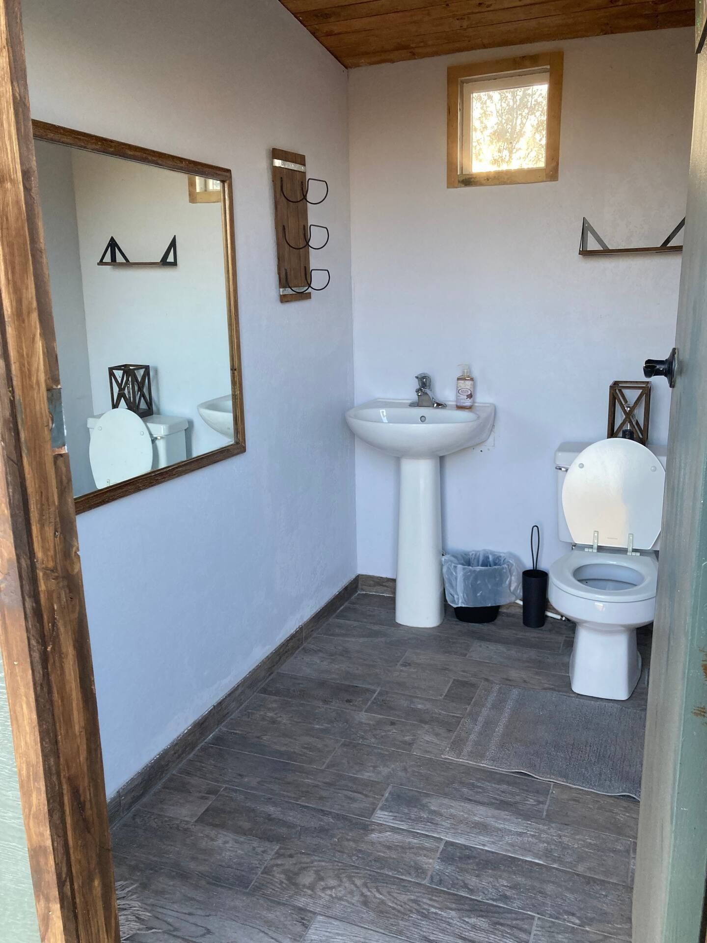 Small bathroom with white walls, a sink, and a toilet. Wooden accents, a mirror, and a small window are also visible.