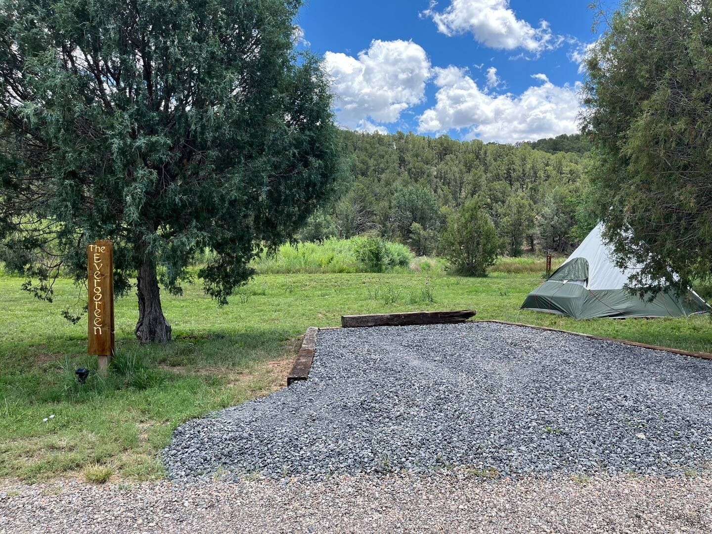 Camping scene: tent, gravel site, trees, sign, hillside, blue sky with clouds.