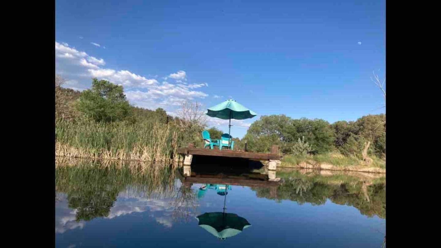 Dock with turquoise umbrella and chairs over calm water reflecting the blue sky and clouds.