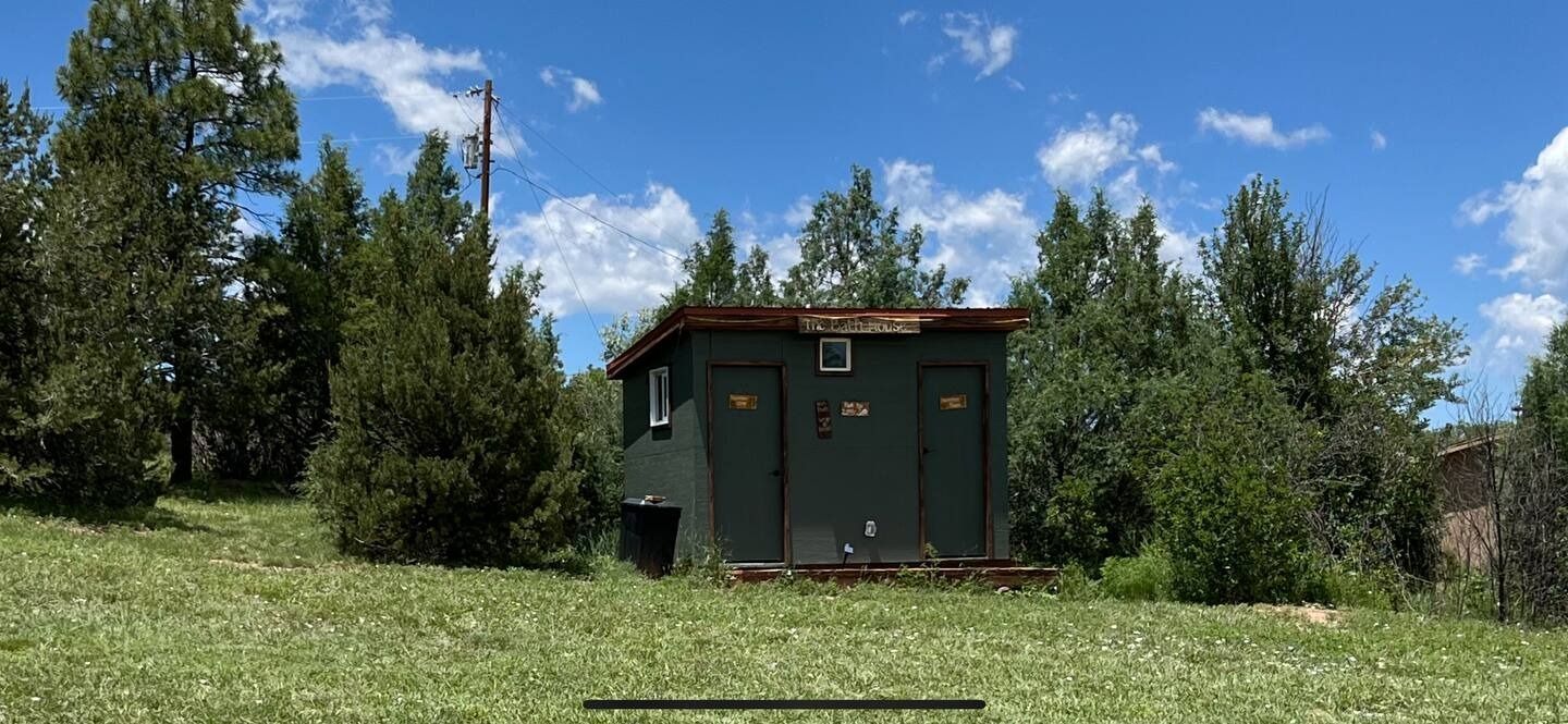 A small, dark green building with two doors and a window set in a grassy field with trees under a blue sky.