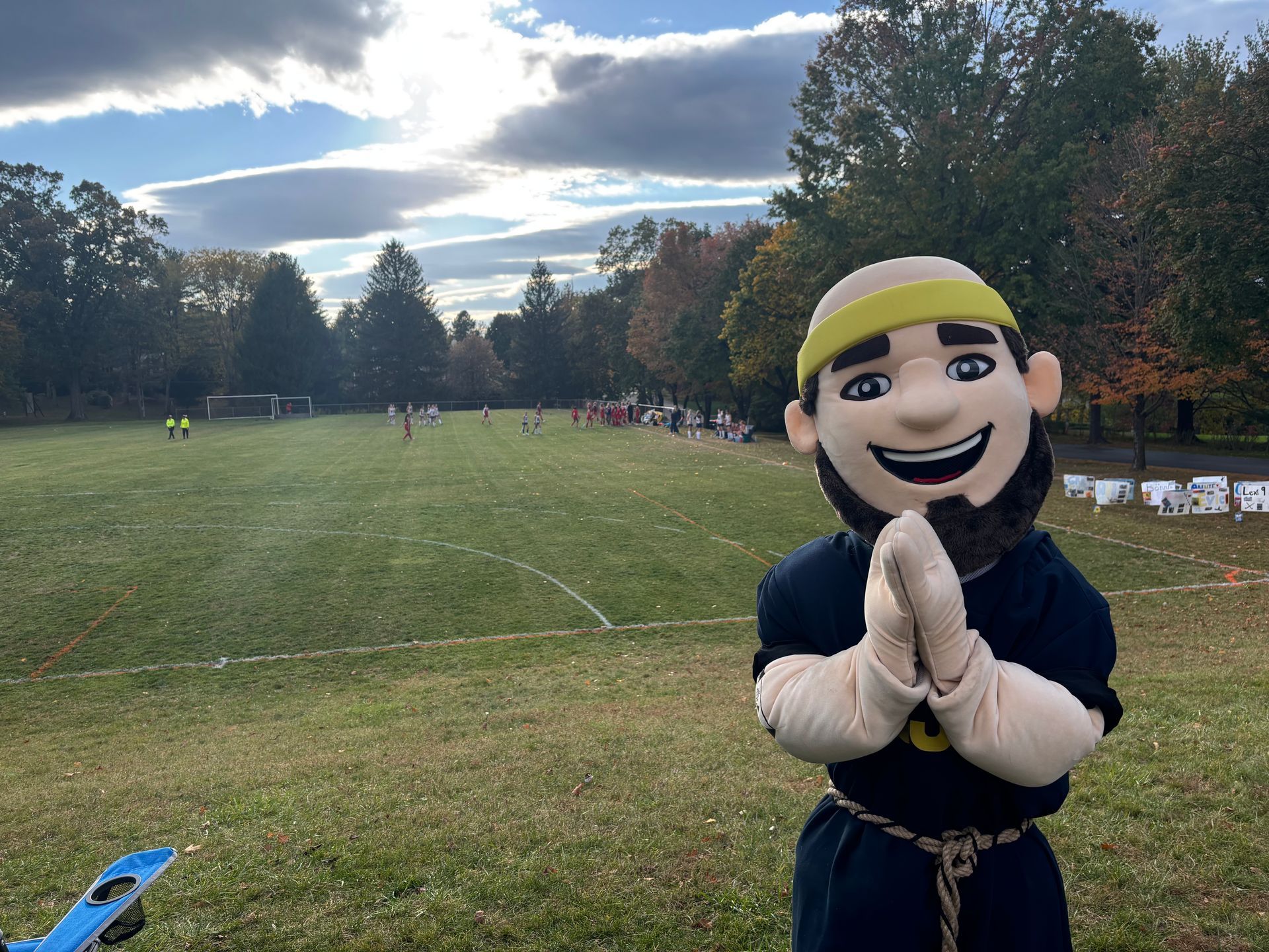 Mascot in black outfit and yellow cap poses in a grassy park under a cloudy sky