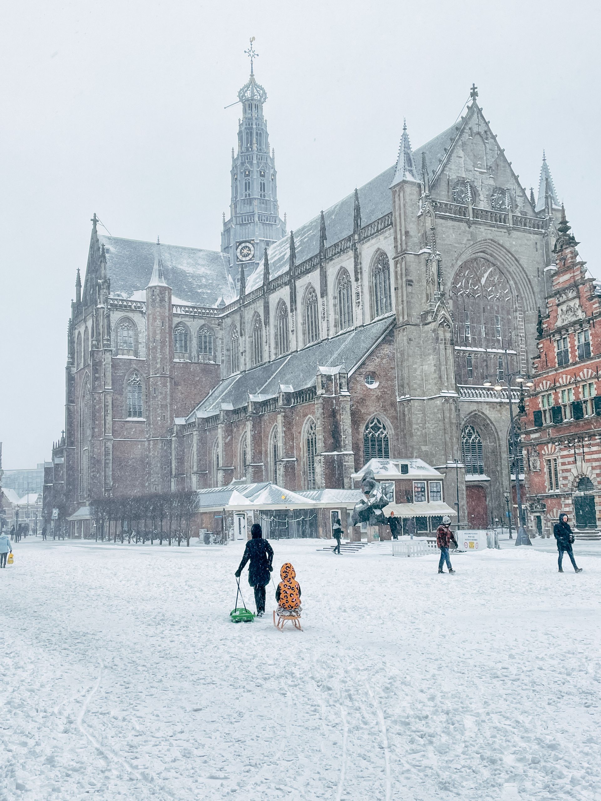 Besneeuwd stadsgezicht met een grote kerk. Iemand trekt een kind op een slee. Andere mensen lopen rond.