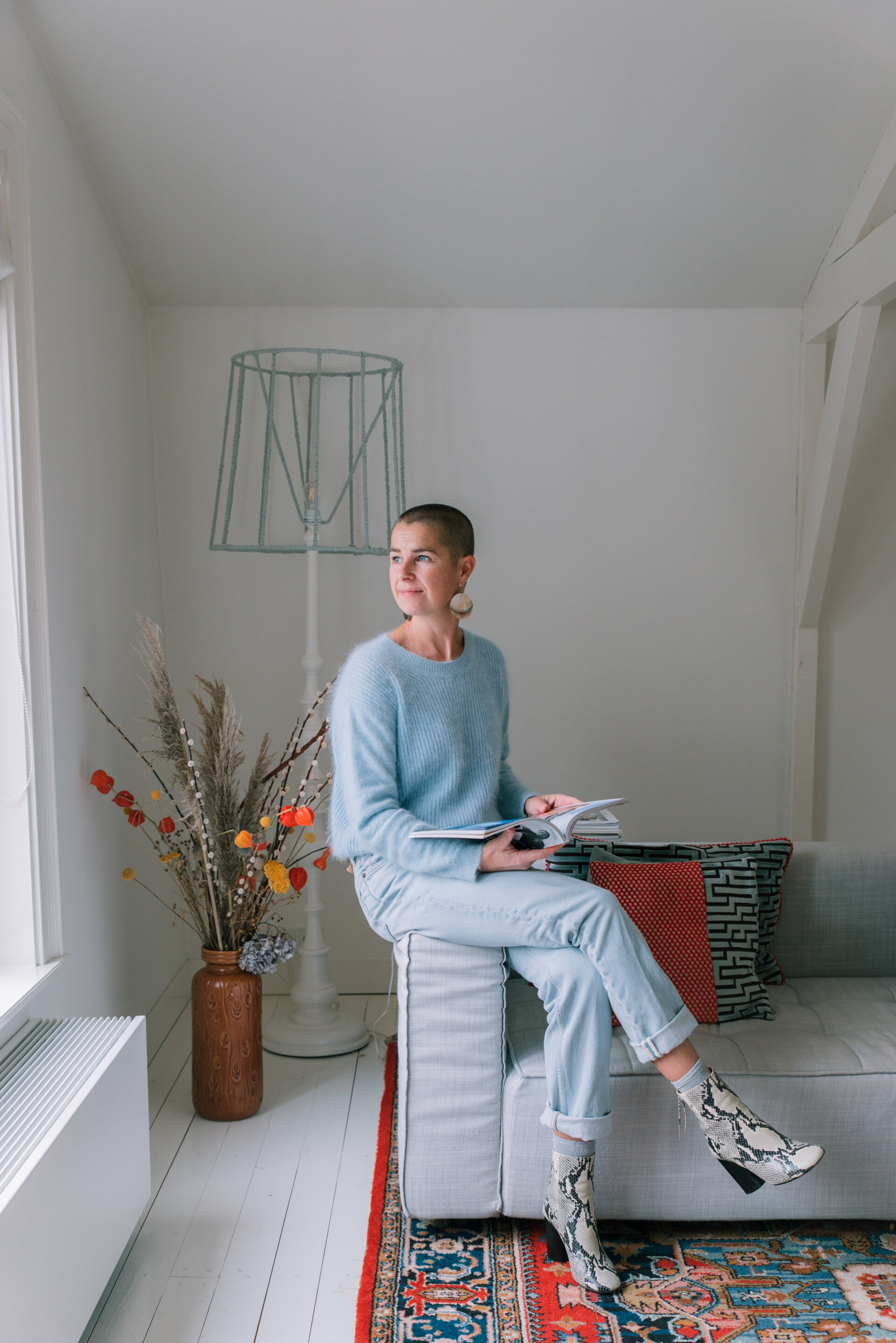 Een vrouw met een kaalgeschoren hoofd zit op de bank, houdt een notitieboekje vast en kijkt uit het raam. Ze draagt een blauwe trui, een spijkerbroek en laarzen van slangenleer.