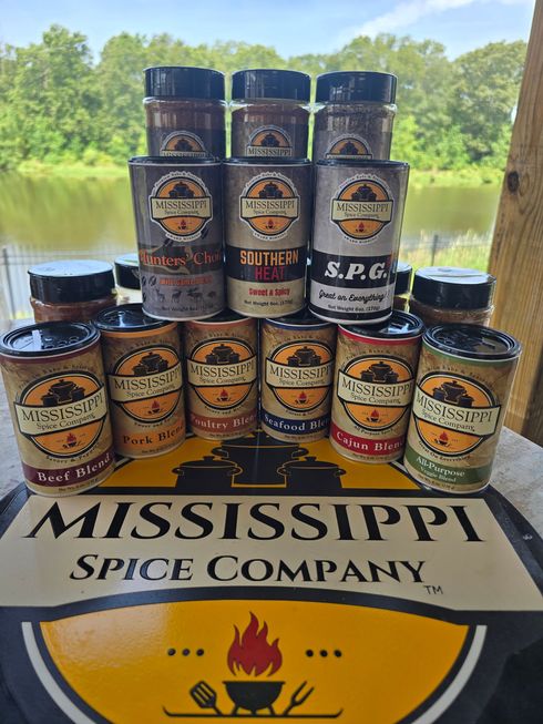 Stacks of Mississippi Spice Company seasoning jars on a branded table outdoors by a lake.