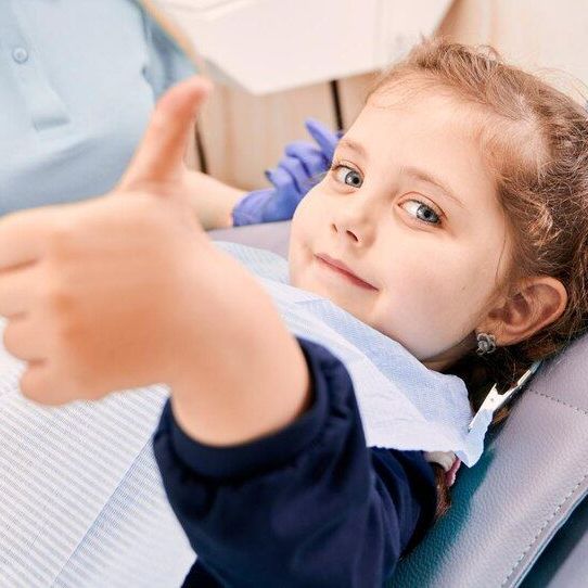 A little girl is giving a thumbs up while sitting in a dental chair.