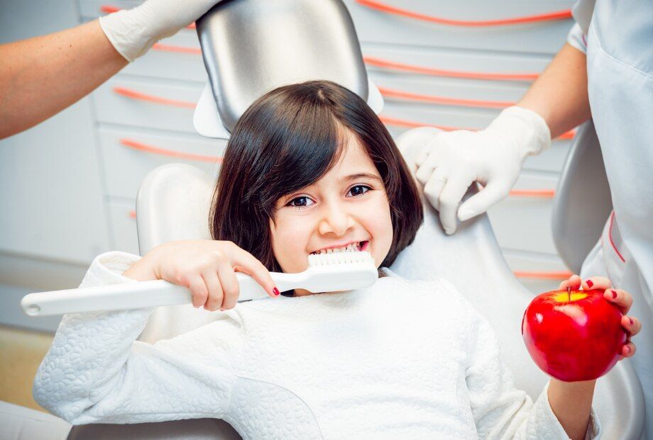 A little girl is brushing her teeth in a dental chair while holding an apple.