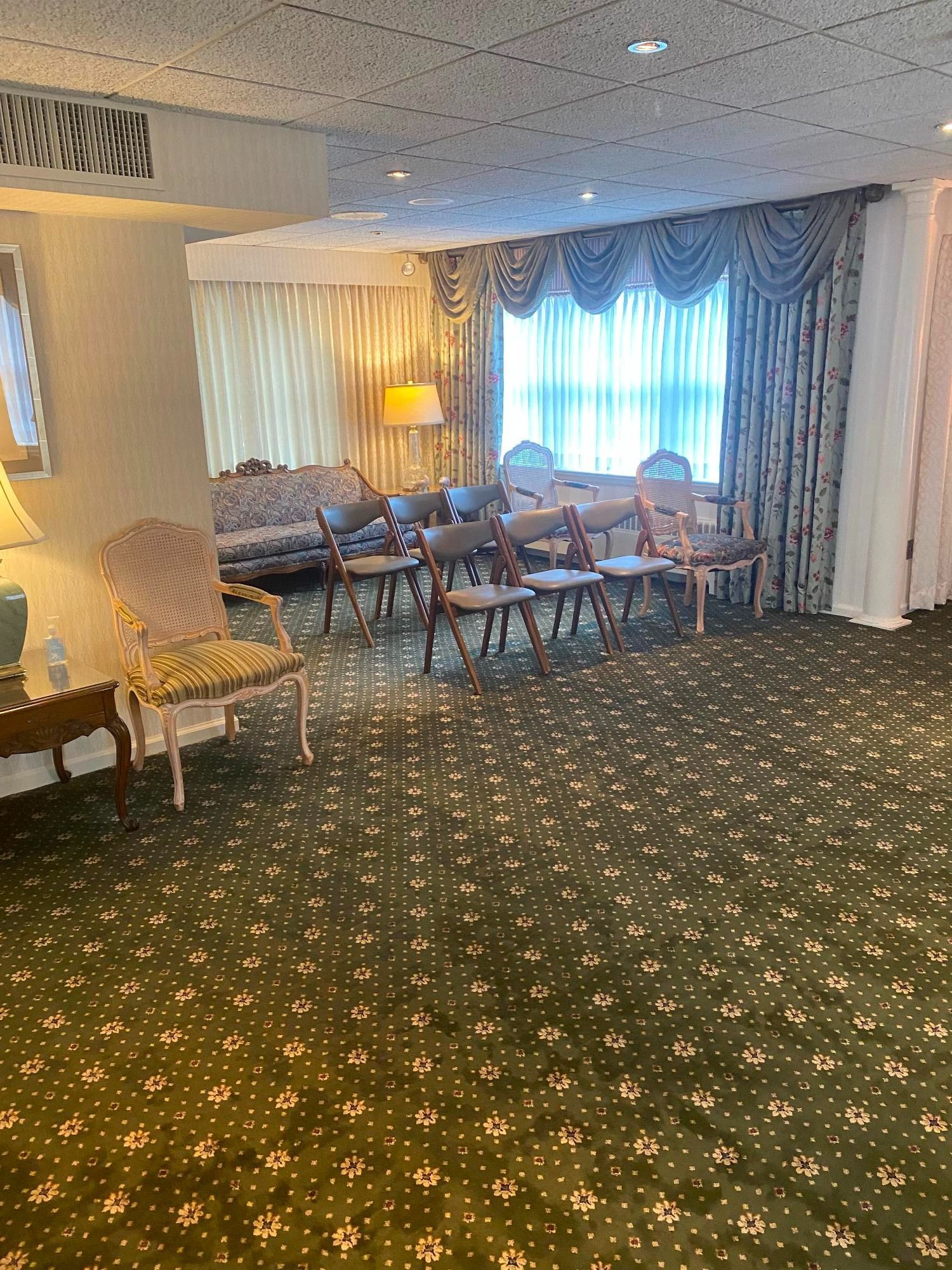 Funeral home interior, green patterned carpet, chairs, sofa, window with draped curtains.