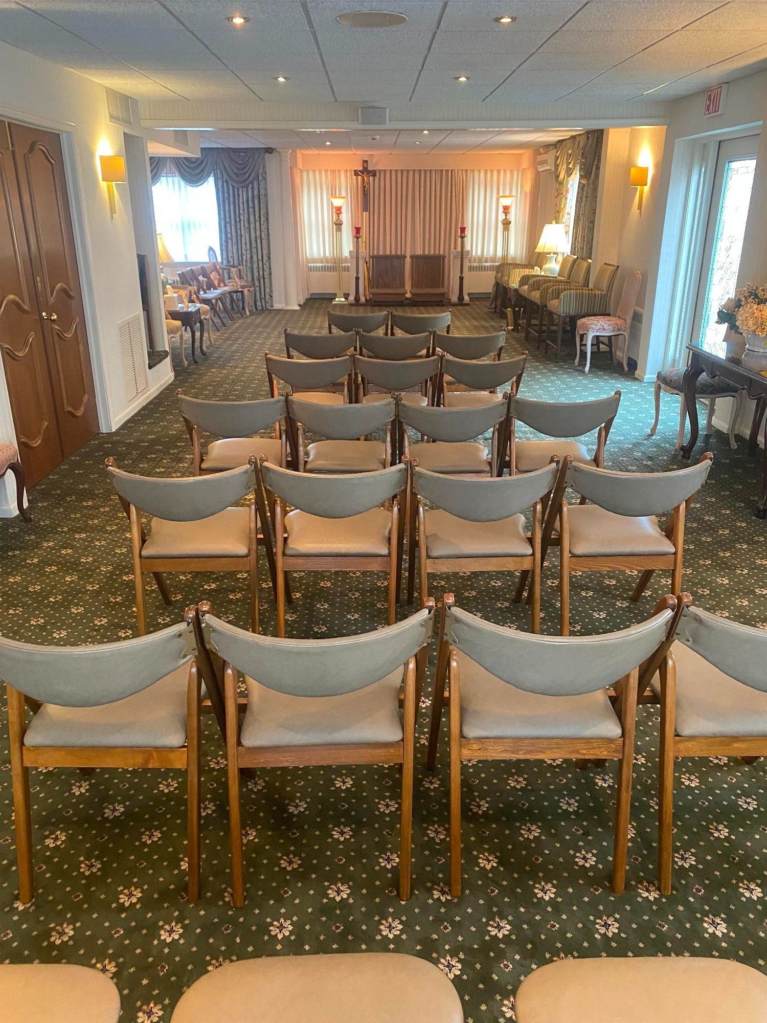 Rows of chairs arranged in a room, likely a chapel. Light green carpet. Neutral colors, with a cross at the end.