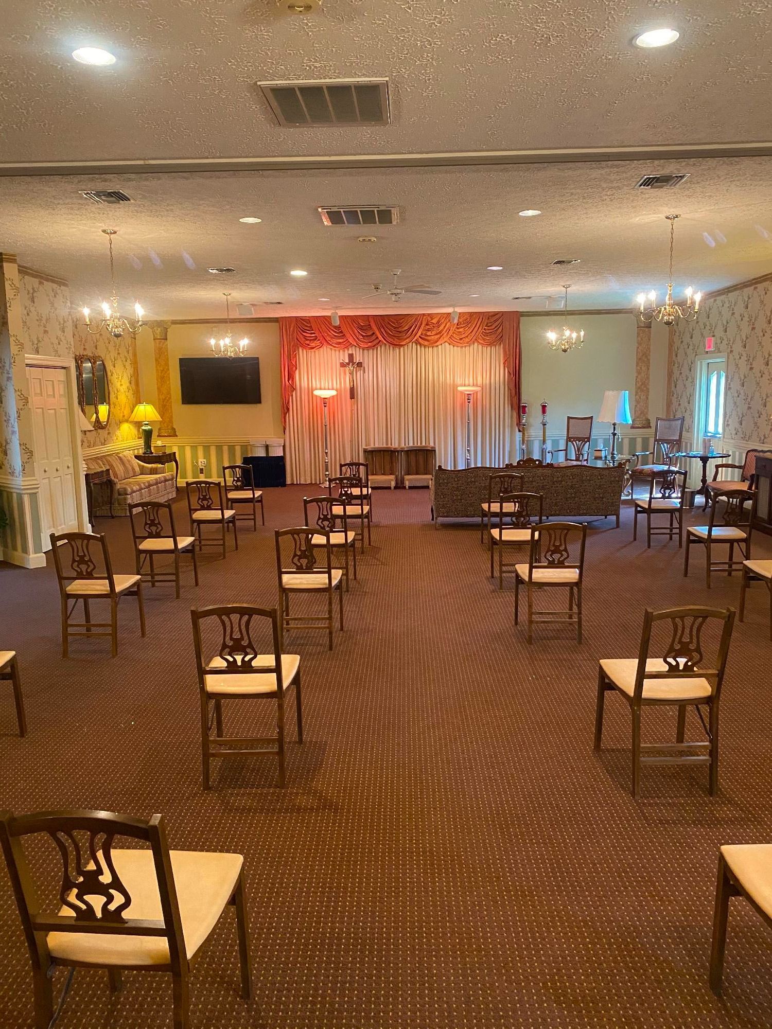 Interior with rows of chairs spaced apart, a stage with a red backdrop, and chandeliers.