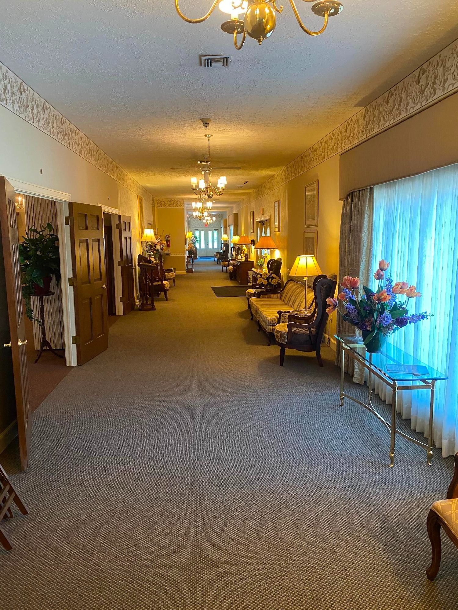 Long hallway with patterned carpet, ornate ceiling lights, and doors on the left, leading to an open area with furniture.