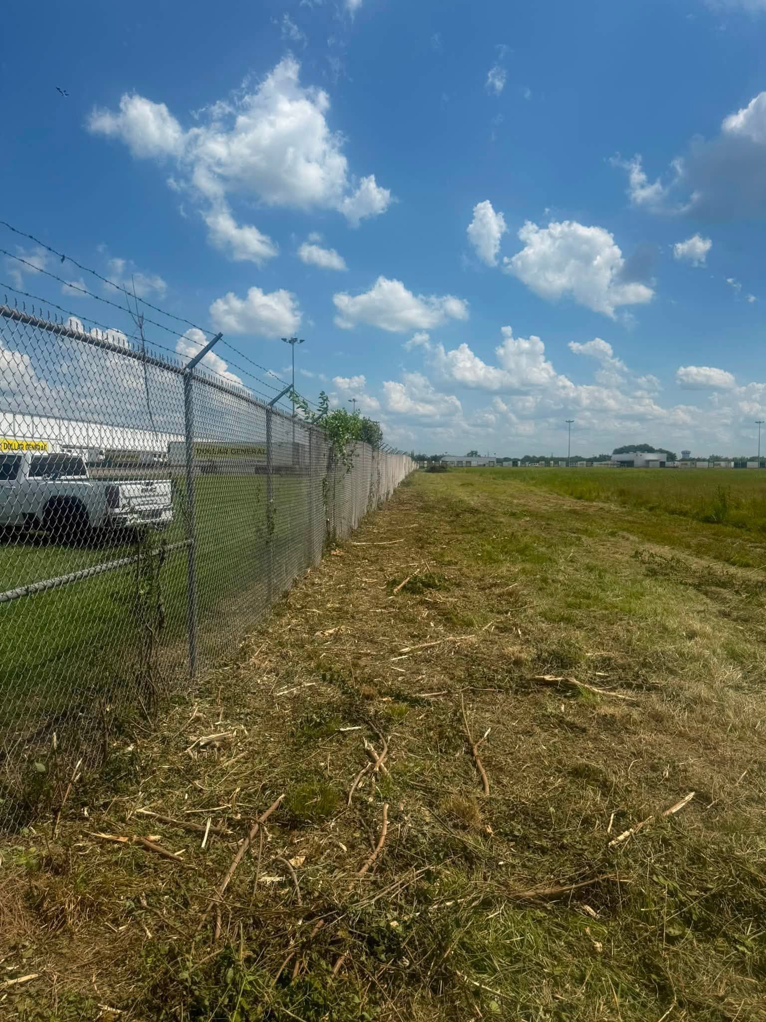 Chain-link fence beside a grassy airfield under a blue sky with scattered clouds