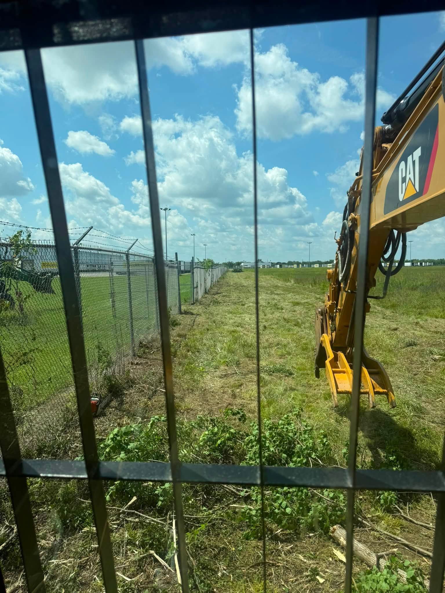 Excavator arm beside a chain-link fence at a grassy construction site under a blue sky.