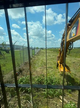 Excavator arm beside a chain-link fence at a grassy construction site under a blue sky.